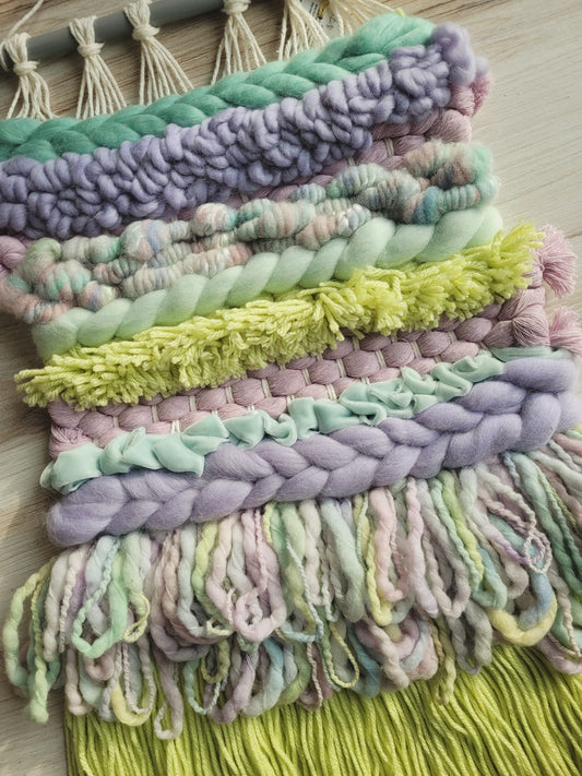 Up close image if a fiber weaving using different sizes and textures of yarn and woving in pastel blues, purples and greens