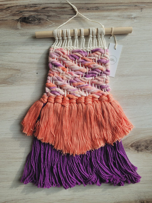 Woven wall hanging on a dowel rod using coral and purple rope featuring a cascading row of coral tassels and then purple yarn fringe. 