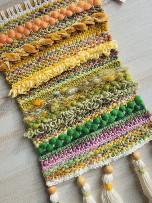 Close up of woven wall hanging made of orange, yellow, greena and purple fibers. There are white tassels with natural wooden beads on the bottom.