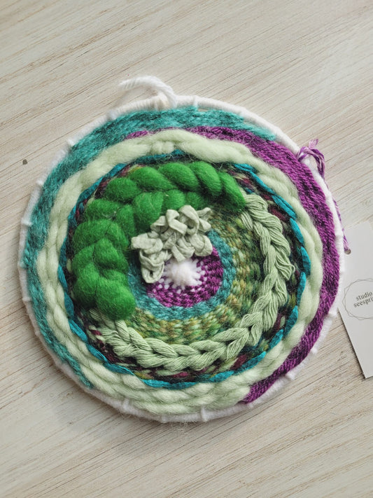 6" round woven wall hanging using a variety of textures and sizes of fibers including roving and yarn in teal, light green, kelly green and purple. 