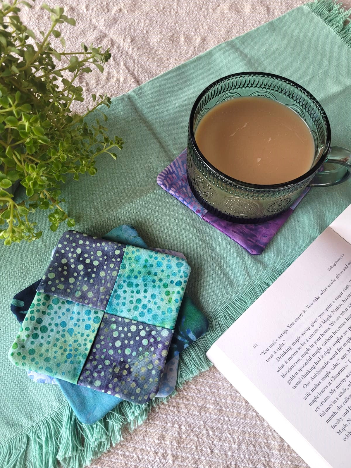 A cup of coffee sits on a purple fabric coaster and a stack of blue fabric coasters sit beside it.