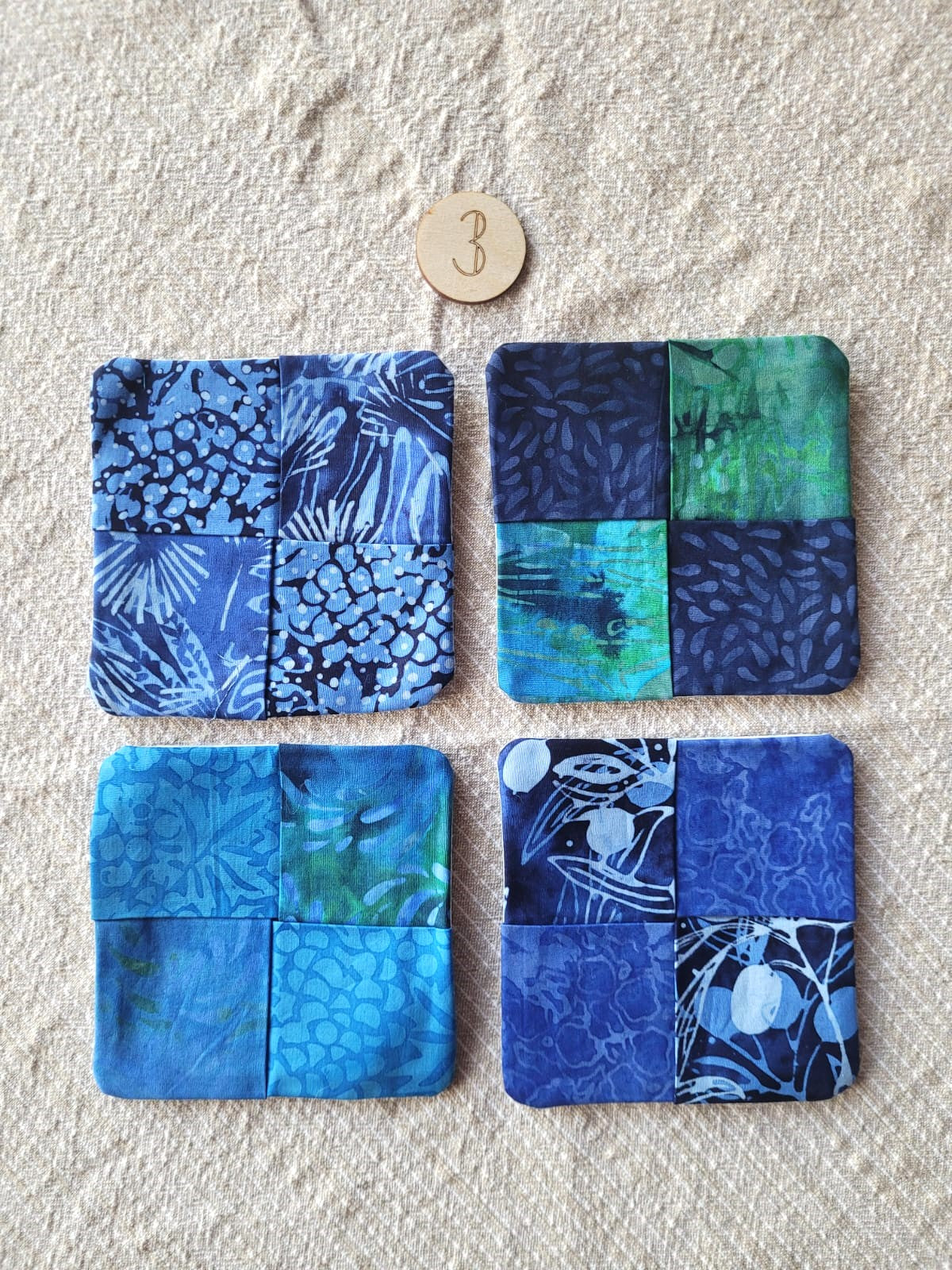 A set of four square fabric coasters made of batik fabrics in blues and greens. Each coaster is different prints. 