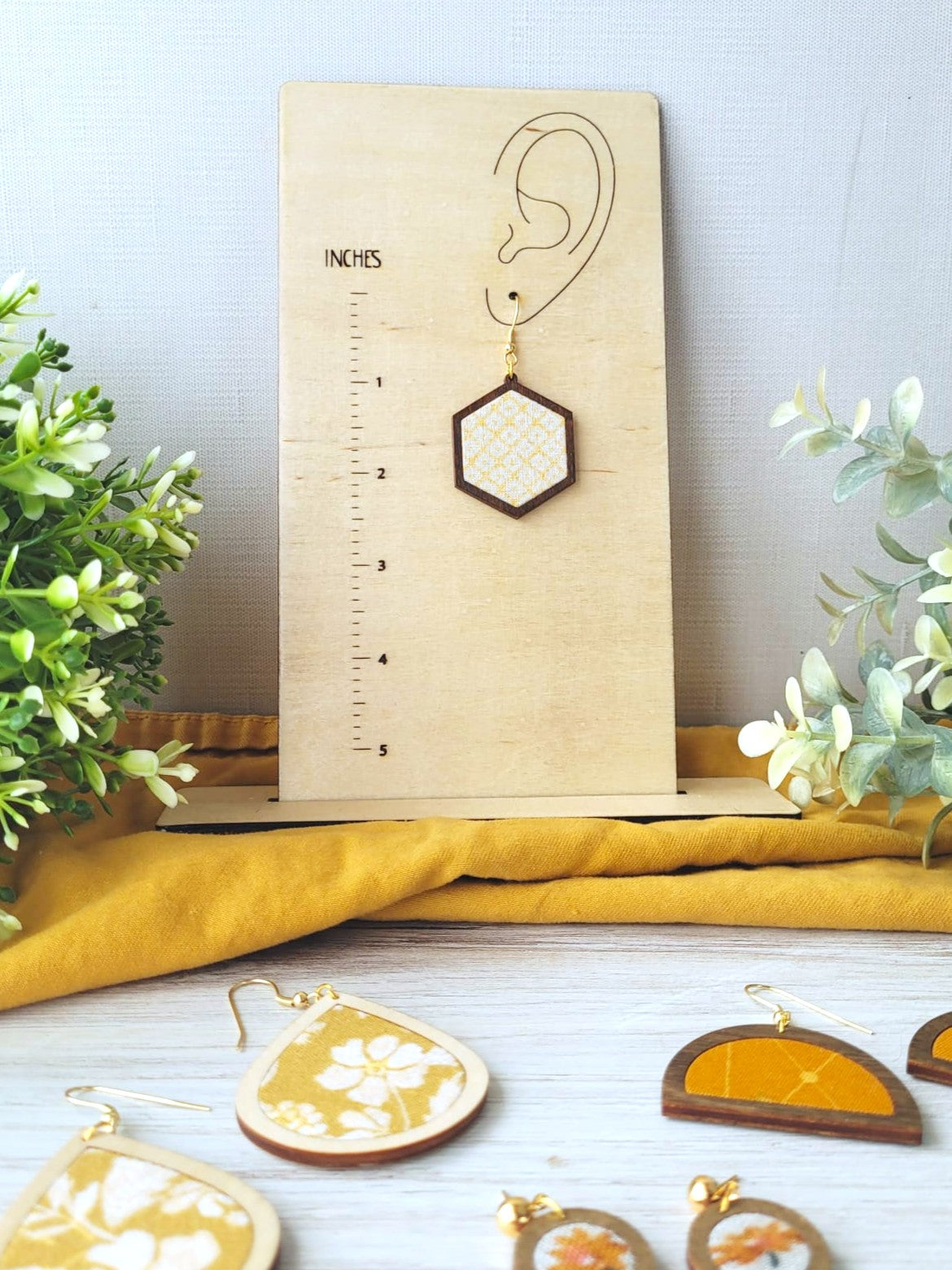 Measurement stand is a rectangular piece of wood that has a ruler printed onto it as well as an ear. The ear has a hole where an earring hook slips through to represent how long the earring hangs from the earlobe. This photo features the stand with an earring on it as well as some greenery and earrings in the foreground. 