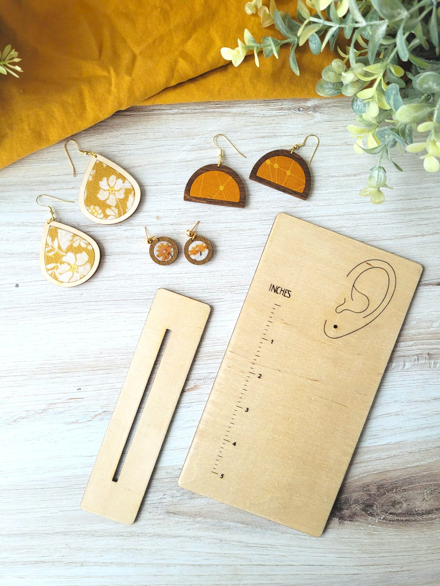 Flatlay of the measurement stand. Measurement stand is a rectangular piece of wood that has a ruler printed onto it as well as an ear. The ear has a hole where an earring hook slips through to represent how long the earring hangs from the earlobe. This photo features the stand with an earring on it as well as some greenery and earrings alongside it. 