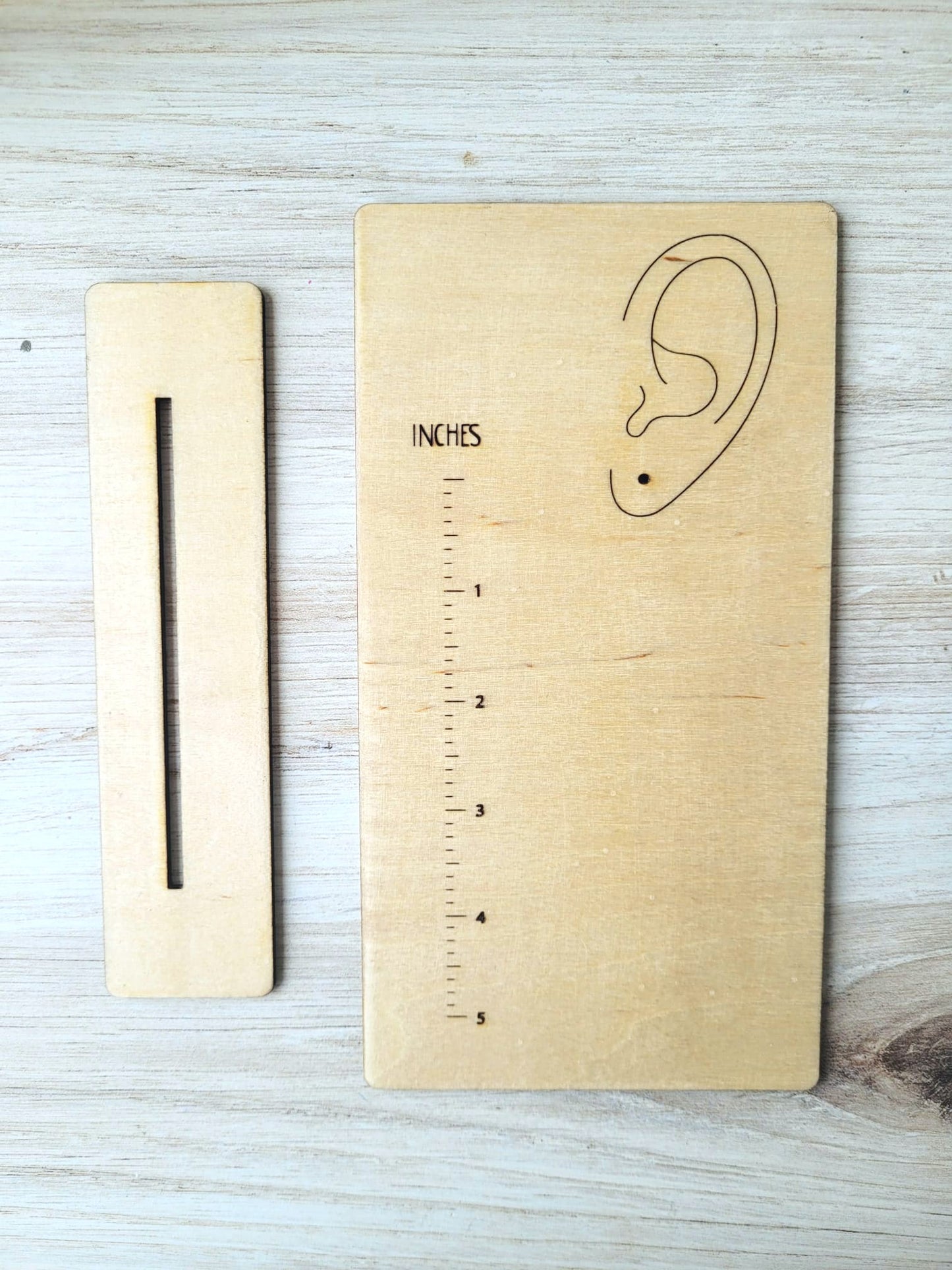 Measurement stand is a rectangular piece of wood that has a rule printed onto it as well as an ear. The ear has a hole where an earring hook slips through to represent how long the earring hangs from the earlobe. 